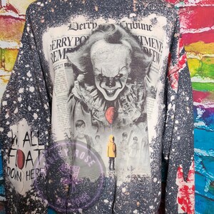 May include: A dark gray sweatshirt with a distressed, speckled design. The front features a newspaper-style graphic of a clown with a red balloon, and the text "Derry Tribune". The left sleeve has the text "WE ALL FLOAT DOWN HERE".