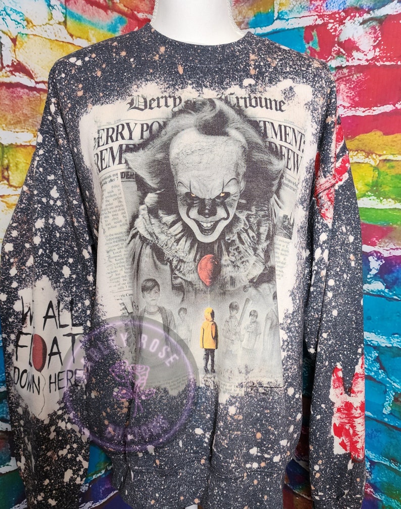 May include: A dark gray sweatshirt with a distressed, speckled design. The front features a newspaper-style graphic of a clown with a red balloon, and the text "Derry Tribune". The left sleeve has the text "WE ALL FLOAT DOWN HERE".