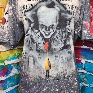 May include: Gray t-shirt featuring a graphic of Pennywise the clown from the movie "It". The design includes the text "Derry Tribune" and a red balloon. The shirt has a speckled, distressed effect.