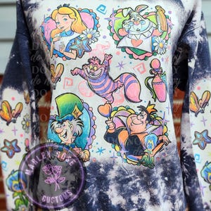 May include: A navy blue sweatshirt with a bleach-splattered design featuring characters from Alice in Wonderland. The characters include Alice, the Mad Hatter, the Cheshire Cat, the Queen of Hearts, and the White Rabbit. The sweatshirt has a crew neck and long sleeves.