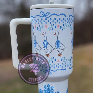 Puede incluir: Taza de viaje de cer&aacute;mica blanca con un dise&ntilde;o floral azul y blanco y tres gansos blancos con cintas azules alrededor del cuello. La taza tiene un asa y una tapa con una pajita.