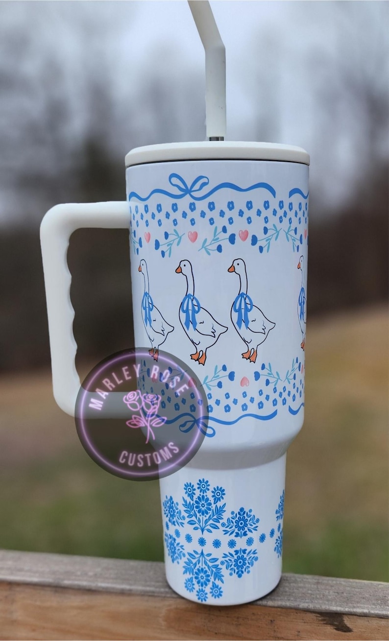 Puede incluir: Taza de viaje de cer&aacute;mica blanca con un dise&ntilde;o floral azul y blanco y tres gansos blancos con cintas azules alrededor del cuello. La taza tiene un asa y una tapa con una pajita.