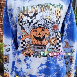 May include: A blue and white tie-dye sweatshirt featuring a graphic print with the text HALLOWEENTOWN, a skeleton, a large pumpkin, a taxi, and a bus stop sign. The sleeves are decorated with smaller pumpkin, witch hat, and star illustrations.