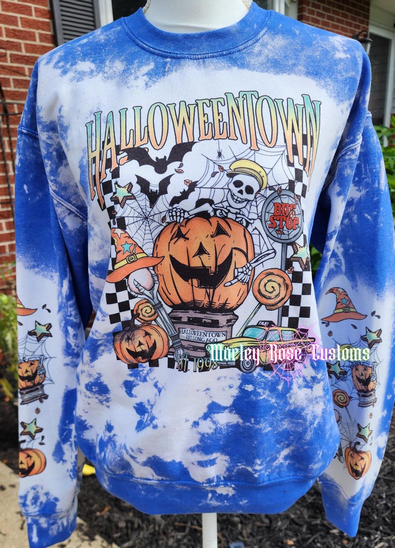 May include: A blue and white tie-dye sweatshirt featuring a graphic print with the text HALLOWEENTOWN, a skeleton, a large pumpkin, a taxi, and a bus stop sign. The sleeves are decorated with smaller pumpkin, witch hat, and star illustrations.