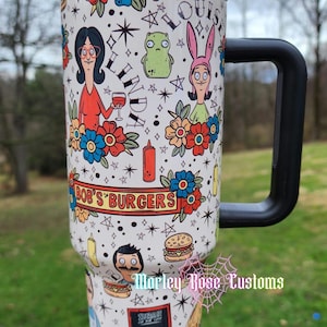 May include: White travel mug featuring Bob's Burgers characters and themed illustrations. The design includes characters like Linda, Louise, and Bob, along with burgers, flowers, and stars. The mug has a black handle and lid.