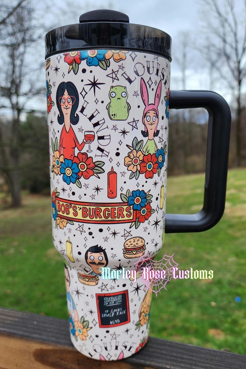 May include: White travel mug featuring Bob's Burgers characters and themed illustrations. The design includes characters like Linda, Louise, and Bob, along with burgers, flowers, and stars. The mug has a black handle and lid.