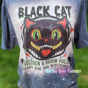 May include: A dark grey short-sleeve t-shirt with a bleached effect. The graphic features a black cat face with green eyes and yellow teeth, surrounded by the text BLACK CAT, CAULDRON & BROOM POLISH, READY FOR THE WITCHING HOUR, and Marley Rose Customs.