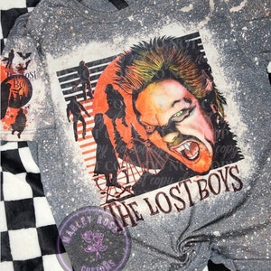 May include: A gray t-shirt with a bleach-splattered design featuring a portrait of a vampire from the movie "The Lost Boys". The vampire has green eyes and fangs, and is surrounded by a red and orange moon. The text "The Lost Boys" is printed in black below the image.