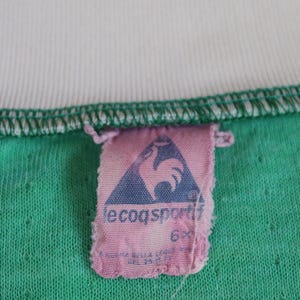 May include: Close-up of a vintage green t-shirt with a white collar. A pink tag with the Le Coq Sportif logo, a blue triangle with a rooster, is attached to the shirt. The shirt has a textured appearance.