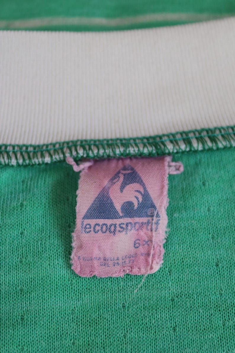 May include: Close-up of a vintage green t-shirt with a white collar. A pink tag with the Le Coq Sportif logo, a blue triangle with a rooster, is attached to the shirt. The shirt has a textured appearance.