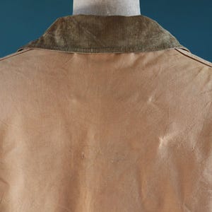 May include: A tan jacket with a corduroy collar is displayed from the back. The jacket's fabric has a slightly worn appearance, with subtle variations in color. The collar is a darker shade of brown, providing a contrast to the jacket's main color.