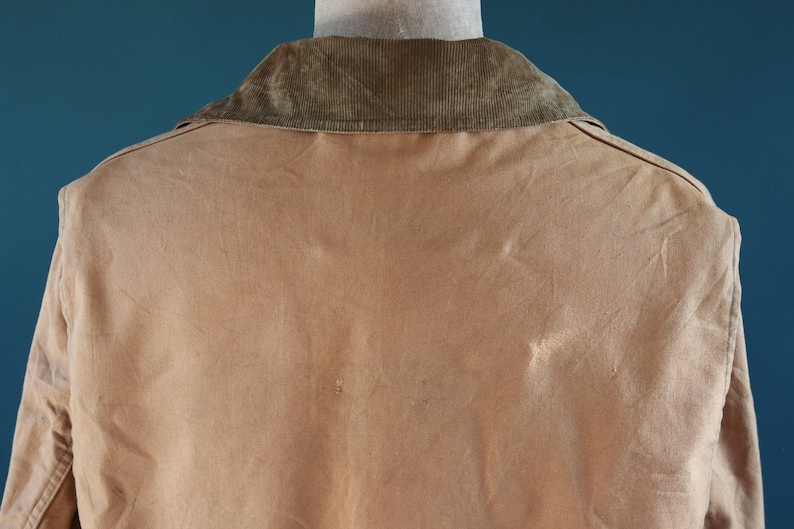 May include: A tan jacket with a corduroy collar is displayed from the back. The jacket's fabric has a slightly worn appearance, with subtle variations in color. The collar is a darker shade of brown, providing a contrast to the jacket's main color.