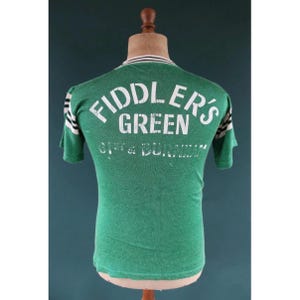 May include: Green t-shirt with the text "FIDDLER'S GREEN" and "CITY & BURNHAM" printed on the back. The shirt has white and black stripes on the sleeves and collar. The shirt is displayed on a mannequin.