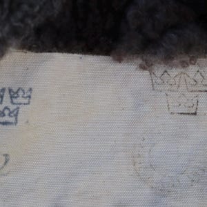May include: Close-up of a light-colored fabric with faded blue crown symbols and the number "2" printed on it. The top of the fabric has a dark, textured material.