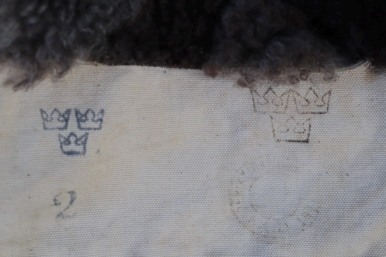 May include: Close-up of a light-colored fabric with faded blue crown symbols and the number "2" printed on it. The top of the fabric has a dark, textured material.