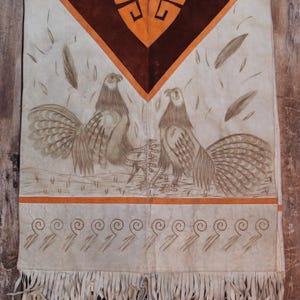 May include: A white leather poncho with brown and orange accents. The poncho features a geometric design at the neck and two stylized birds with long tails. The poncho has a fringe border.