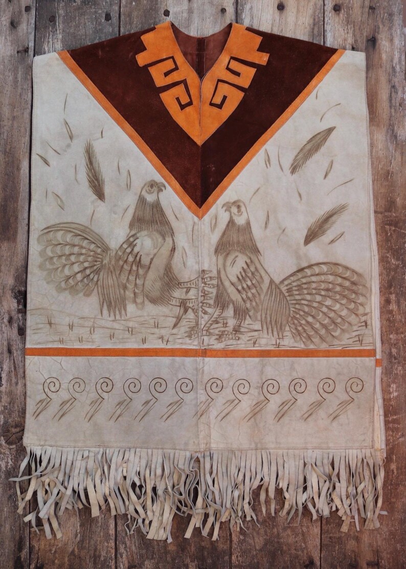 May include: A white leather poncho with brown and orange accents. The poncho features a geometric design at the neck and two stylized birds with long tails. The poncho has a fringe border.