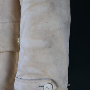 May include: Close-up of a section of a light beige canvas jacket sleeve. The sleeve features a buttoned cuff with a white button and a small loop. The fabric shows signs of wear and staining, suggesting a vintage or workwear aesthetic.