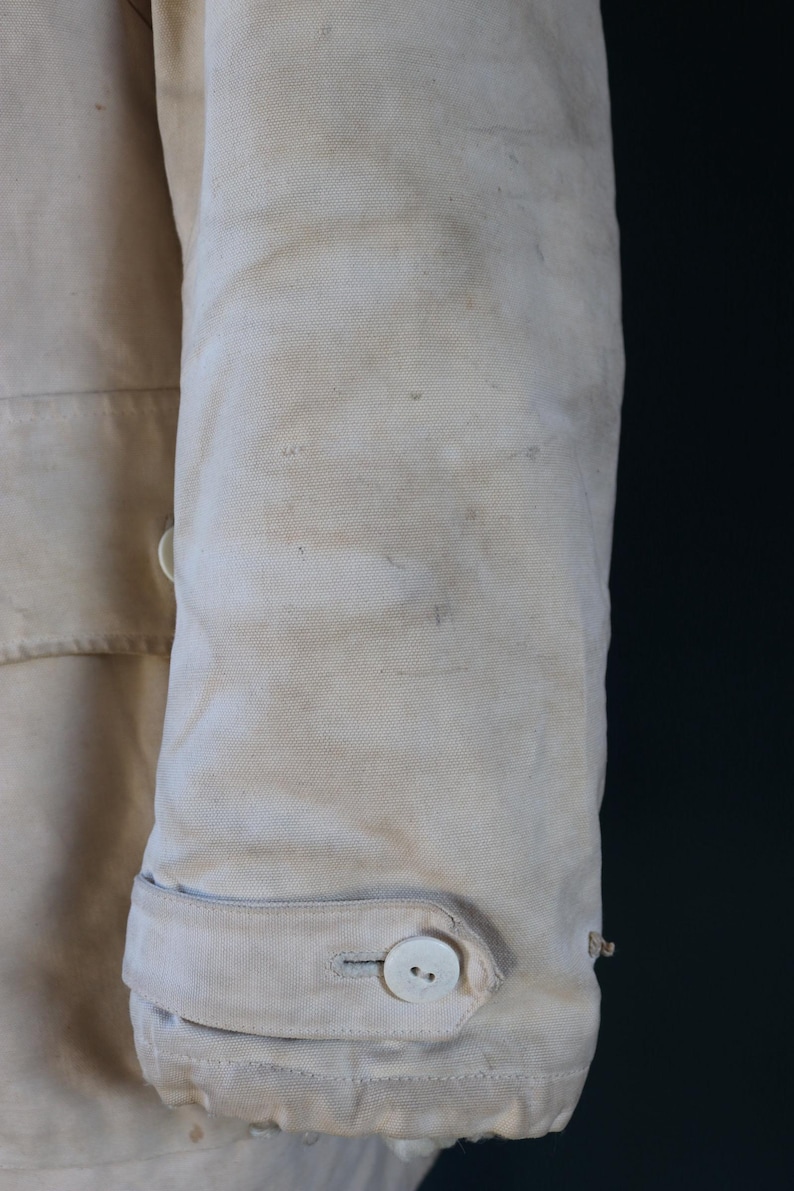 May include: Close-up of a section of a light beige canvas jacket sleeve. The sleeve features a buttoned cuff with a white button and a small loop. The fabric shows signs of wear and staining, suggesting a vintage or workwear aesthetic.