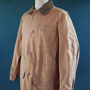 May include: A tan work jacket with a corduroy collar, button-up front, and multiple pockets. The jacket is displayed on a mannequin. The jacket has a classic, utilitarian design.