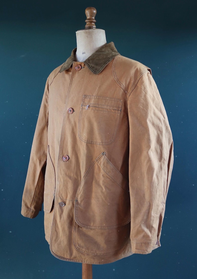 May include: A tan work jacket with a corduroy collar, button-up front, and multiple pockets. The jacket is displayed on a mannequin. The jacket has a classic, utilitarian design.