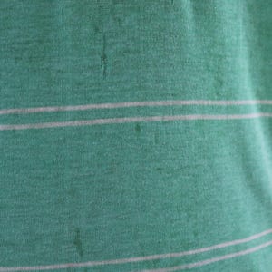 May include: Close-up of a green fabric with two horizontal white stripes. The fabric has a textured appearance and a small white tag at the top. The overall color is a muted green.