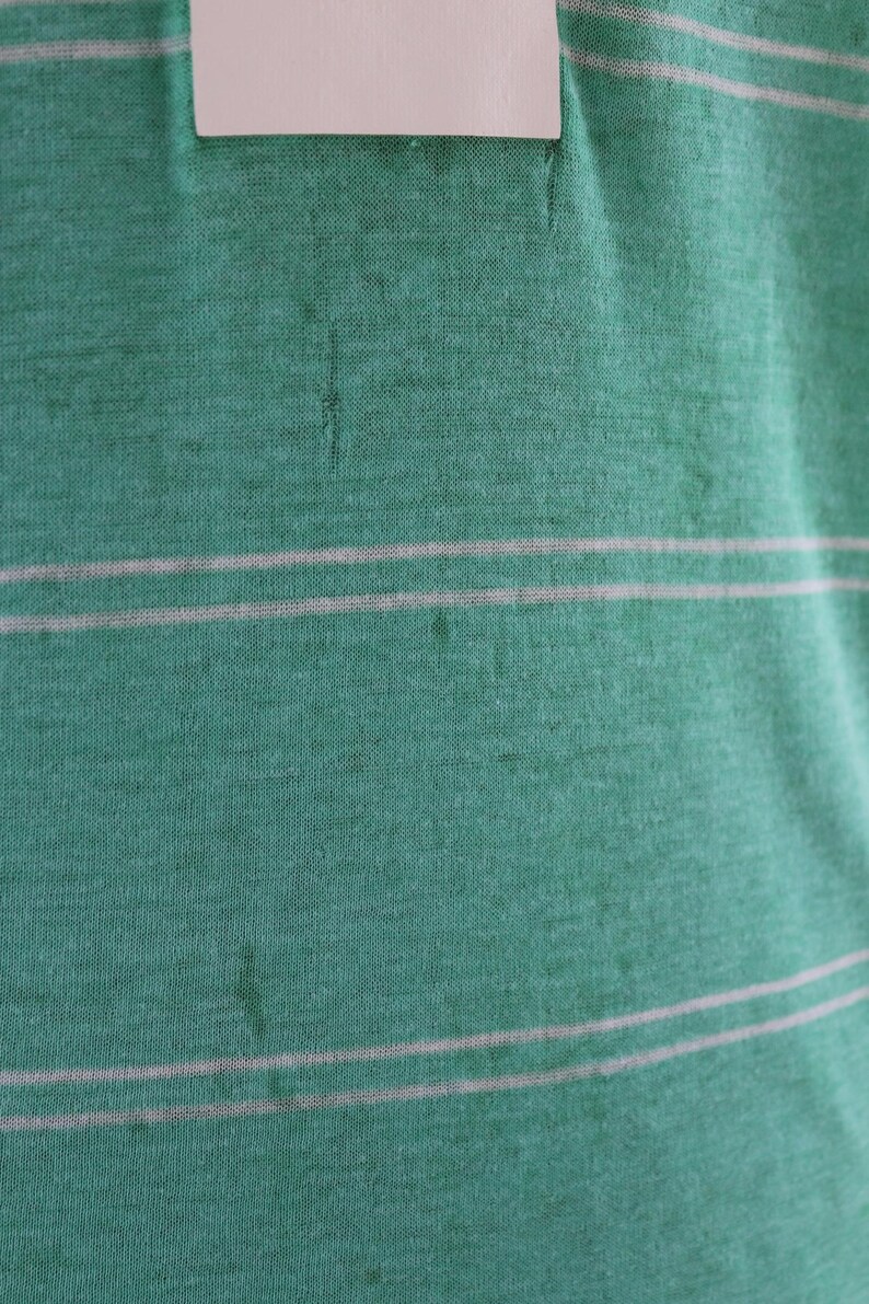 May include: Close-up of a green fabric with two horizontal white stripes. The fabric has a textured appearance and a small white tag at the top. The overall color is a muted green.