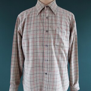 May include: A long-sleeved button-down shirt in a beige and gray plaid pattern with red and black accents. The shirt has a classic collar, a chest pocket, and buttoned cuffs. The shirt is displayed on a wooden mannequin.