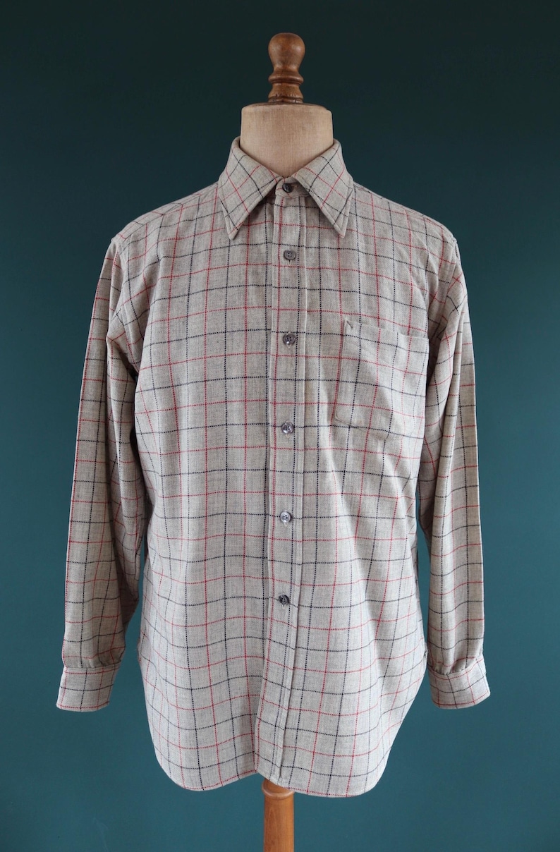 May include: A long-sleeved button-down shirt in a beige and gray plaid pattern with red and black accents. The shirt has a classic collar, a chest pocket, and buttoned cuffs. The shirt is displayed on a wooden mannequin.