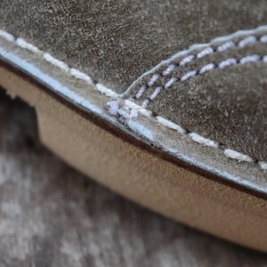 May include: Close-up of a section of a shoe, showcasing the textured, gray suede material. White stitching accents the seams, with a light blue trim. The sole is a light brown color.