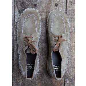 May include: A pair of olive green suede shoes with brown laces, stitched detailing, and a rounded toe design. The shoes have a classic, casual style, suitable for everyday wear. The soles are a light tan color.