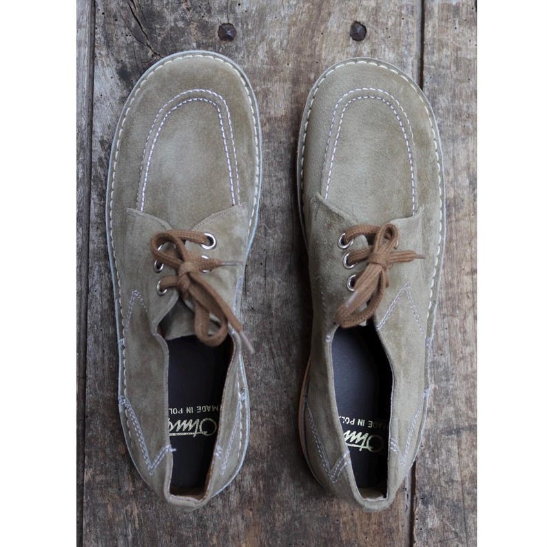 May include: A pair of olive green suede shoes with brown laces, stitched detailing, and a rounded toe design. The shoes have a classic, casual style, suitable for everyday wear. The soles are a light tan color.