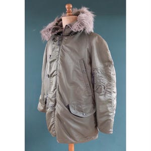 May include: An olive green parka with a faux fur-lined hood. The jacket has multiple pockets, a zip closure, and button details. The parka is displayed on a wooden mannequin.