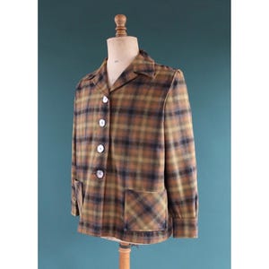 May include: A vintage plaid jacket in shades of brown, olive green, and black, displayed on a mannequin. The jacket has a classic collar, three white buttons, two front pockets, and long sleeves. The fabric has a textured appearance.