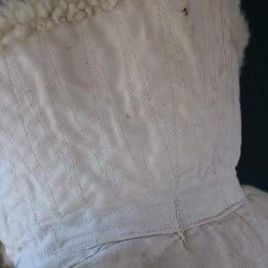 May include: Close-up of a cream-colored hood with a fluffy, off-white trim. The hood is made of a textured fabric with vertical stitching details. The trim appears to be a soft, wool-like material.