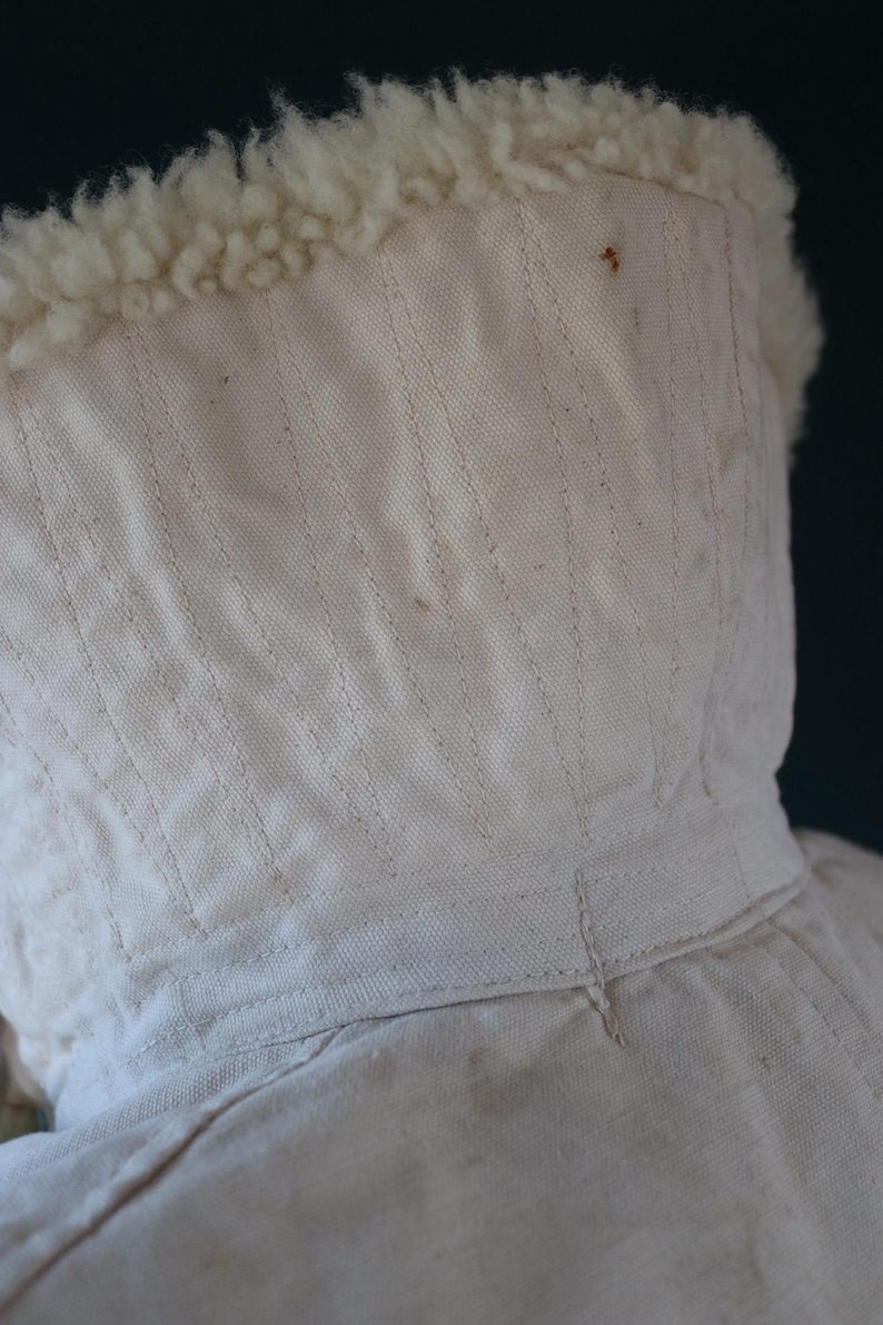 May include: Close-up of a cream-colored hood with a fluffy, off-white trim. The hood is made of a textured fabric with vertical stitching details. The trim appears to be a soft, wool-like material.