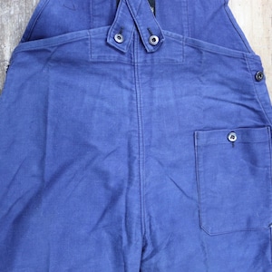 May include: Blue denim dungarees with a single back pocket and two button fastenings on the straps.
