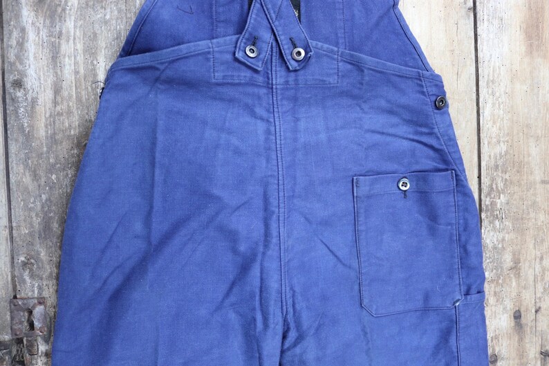 May include: Blue denim dungarees with a single back pocket and two button fastenings on the straps.