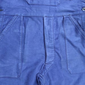 May include: A pair of blue denim dungarees with two large pockets on the front. The dungarees have a button fastening at the waist.