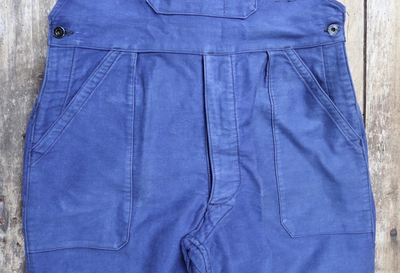 May include: A pair of blue denim dungarees with two large pockets on the front. The dungarees have a button fastening at the waist.