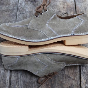 May include: A pair of olive green suede shoes with light brown soles and white stitching. The shoes have brown laces and a classic derby design. The shoes are displayed on a wooden surface.