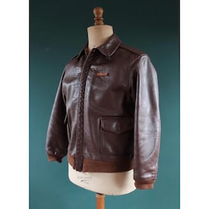 May include: A brown leather bomber jacket with a collared neckline, two front pockets, and a brown ribbed hem and cuffs. The jacket is displayed on a mannequin. A small patch is visible on the chest.