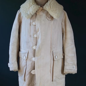 May include: Cream-colored vintage coat with a sheepskin collar. The coat features a button-up front, two front pockets, and buttoned cuffs. The coat is displayed on a wooden mannequin.