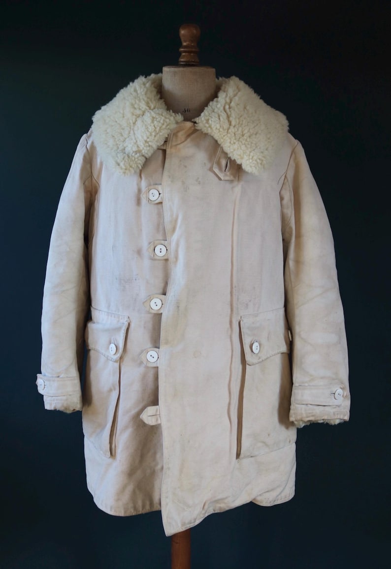 May include: Cream-colored vintage coat with a sheepskin collar. The coat features a button-up front, two front pockets, and buttoned cuffs. The coat is displayed on a wooden mannequin.