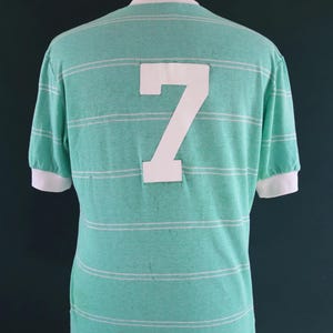 May include: A vintage-style, mint green t-shirt with horizontal white stripes. The back of the shirt features a large white number 7. The short sleeves and collar have white trim. The shirt is displayed on a mannequin.