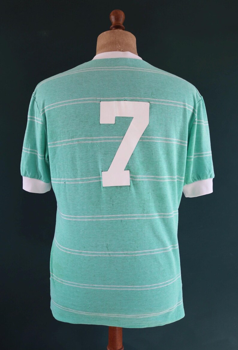 May include: A vintage-style, mint green t-shirt with horizontal white stripes. The back of the shirt features a large white number 7. The short sleeves and collar have white trim. The shirt is displayed on a mannequin.