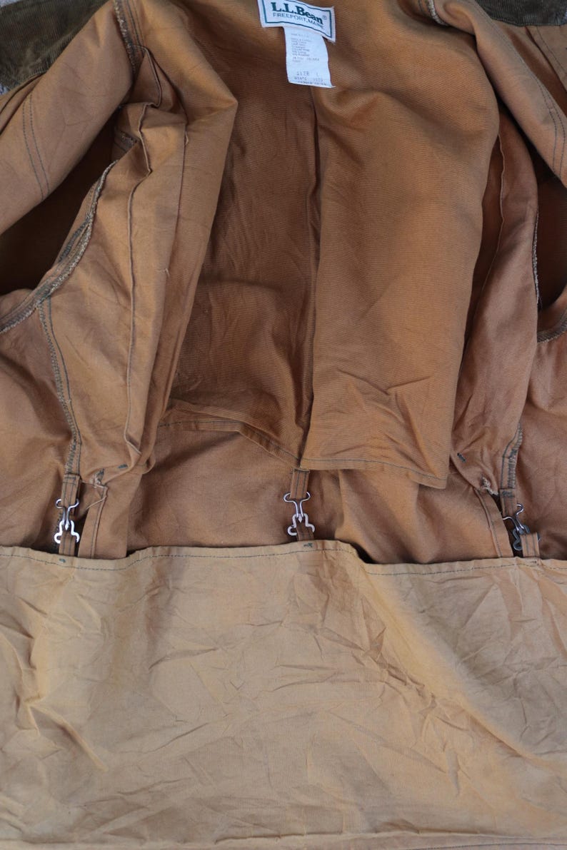 May include: Close-up of the interior of a brown canvas jacket. The jacket has a light brown lining and metal clasps. The L.L.Bean label is visible at the top. The jacket appears to be well-worn.