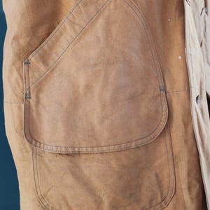 May include: Close-up of a tan work jacket with a large, curved pocket. The jacket has visible stitching and a buttoned front. The fabric appears worn, suggesting a vintage or well-used item. The pocket is a prominent feature.
