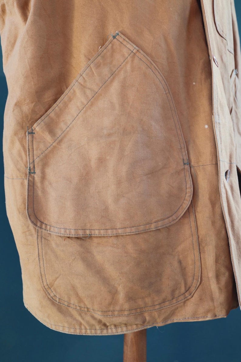 May include: Close-up of a tan work jacket with a large, curved pocket. The jacket has visible stitching and a buttoned front. The fabric appears worn, suggesting a vintage or well-used item. The pocket is a prominent feature.