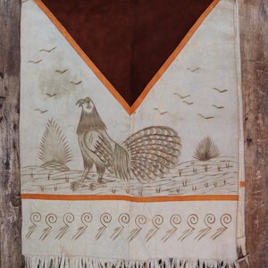 May include: A white leather poncho with a brown suede panel and orange trim. The poncho features a painted design of a bird with a long tail and a fringe border.